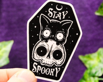 Könnte beinhalten: Ein sargförmiger Aufkleber mit einer schwarzen Katze, die auf einem weißen menschlichen Schädel sitzt, vor einem schwarzen Hintergrund mit weißen Sternen. Der Text STAY SPOOKY ist in weißen Buchstaben gedruckt. Das Design ist weiß umrandet.