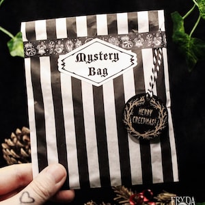 May include: A black and white striped mystery bag with a label reading "Mystery Bag". A round tag with "Merry Creepmas!" hangs from the bag. The bag is held by a hand with a heart tattoo. The overall theme is gothic.
