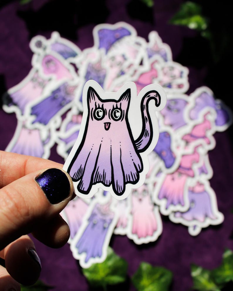 May include: A sticker of a purple and pink ghost cat with big eyes and a wide smile. The sticker is on a purple background with other stickers of ghosts and witches.