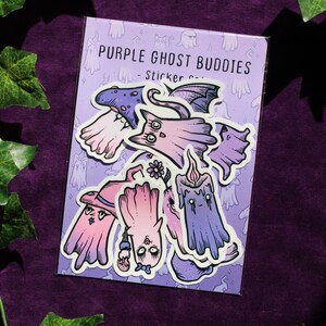 K&ouml;nnte beinhalten: Eine Packung Aufkleber mit verschiedenen lila Geister-Designs. Die Aufkleber zeigen Geister mit H&uuml;ten, Fl&uuml;geln und anderem Zubeh&ouml;r. Der Text "PURPLE GHOST BUDDIES" ist auf der Verpackung aufgedruckt. Die Aufkleber befinden sich auf einem lila Hintergrund.