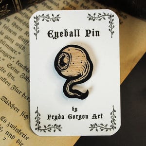 Könnte beinhalten: Holzstift in Form eines Augapfels mit schwarzem Umriss und brauner Holzmaserung. Der Stift befindet sich auf einer weißen Karte mit dem Text "Eyeball Pin" und "by Fryda Gorgon Art".