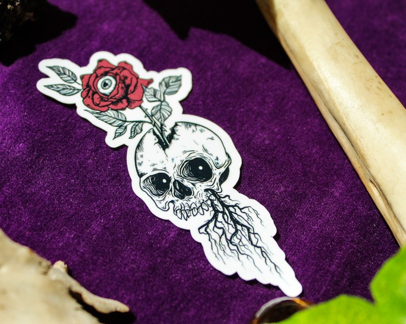 May include: A die-cut sticker featuring a black and white line art illustration of a human skull with a red rose growing out of the top. The rose centre contains a realistic eye, and dark, spindly roots extend from the bottom of the skull jaw.