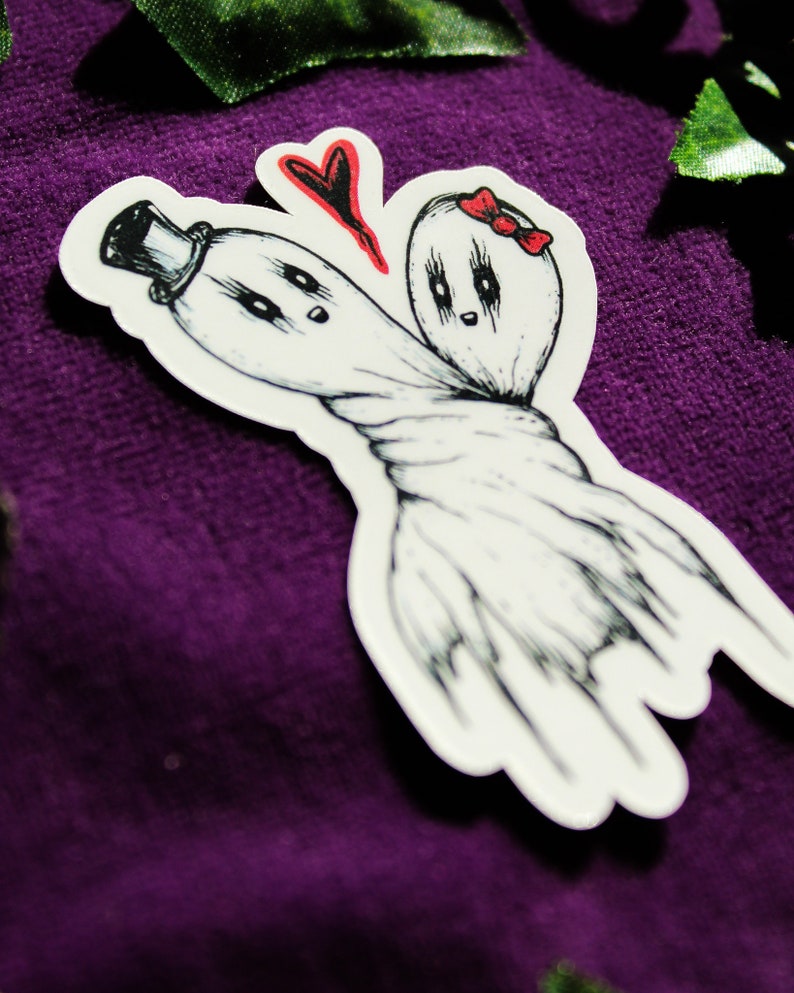 Może przedstawiać: Wykrawana naklejka przedstawiająca dwa białe duszki z czarnymi konturami wykonanymi tuszem. Jeden duszek ma czarny cylinder, drugi czerwoną kokardkę. Są połączone w środkowej części, a nad nimi znajduje się czerwone serce. Styl rysunku przypomina szkic.