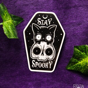May include: A black and white sticker featuring a cat and skull design. The cat sits above the skull, with the words "STAY SPOOKY" in white. The sticker is coffin-shaped and set against a purple background.