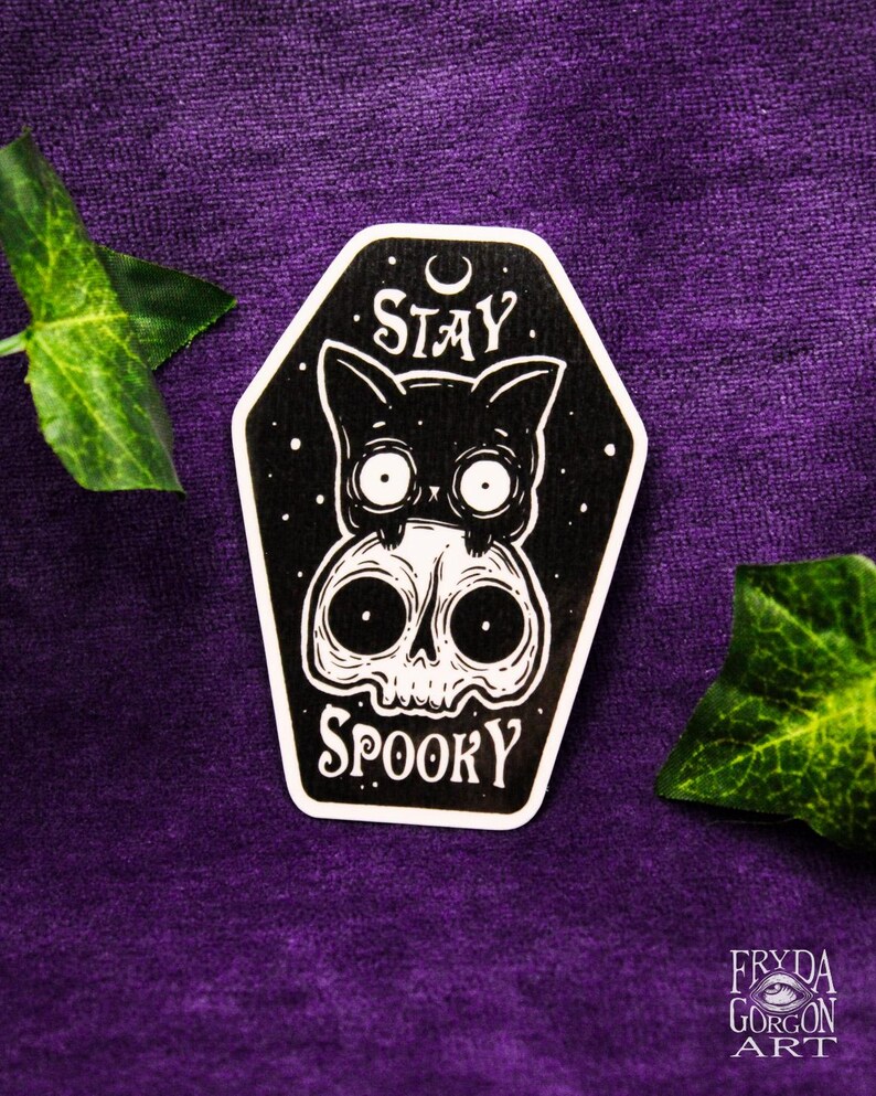May include: A black and white sticker featuring a cat and skull design. The cat sits above the skull, with the words "STAY SPOOKY" in white. The sticker is coffin-shaped and set against a purple background.