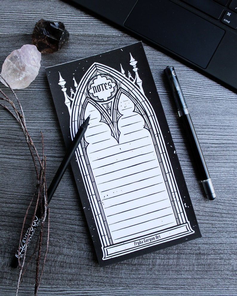 May include: Black and white gothic style notepad with a decorative archway design. The notepad has lined pages and the text "NOTES" in a circle at the top. The bottom of the notepad has the text "Fryda Gorgon Art".