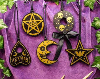 May include: Five black and gold hanging ornaments featuring a pentagram with an eye, a skull wreath with a black ribbon bow, a planchette shape with the text HEXMAS, a crescent moon with a face, and a star with a bat silhouette in the center.