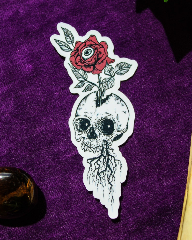 May include: A die-cut sticker featuring a black and white line art illustration of a human skull with roots growing from the jaw. A red rose with a realistic human eye in the centre of the petals emerges from the top of the cracked skull.