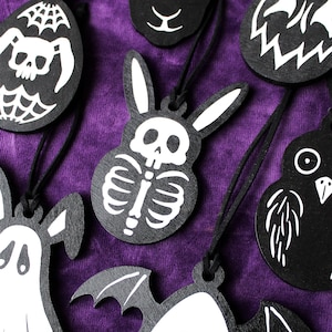 Può includere: Un set di dieci decorazioni di Halloween in legno nero con disegni bianchi. Le decorazioni presentano una varietà di personaggi spettrali, tra cui un fantasma, un teschio, un pipistrello e una zucca.