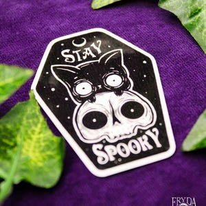 May include: A black and white sticker featuring a cat on a skull, with the words "Stay Spooky". The sticker is set against a purple background with green foliage.