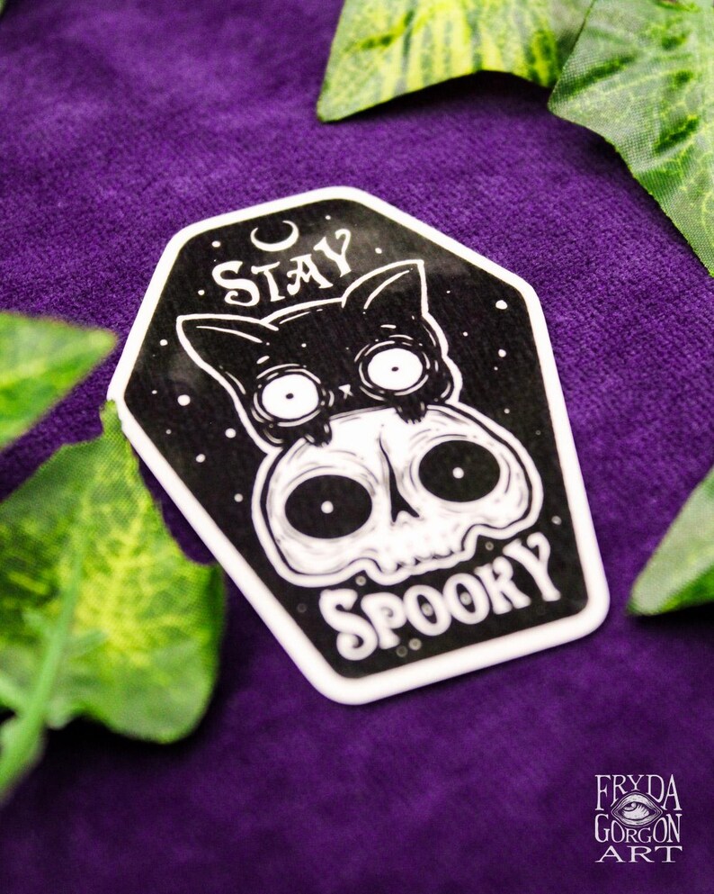 May include: A black and white sticker featuring a cat on a skull, with the words "Stay Spooky". The sticker is set against a purple background with green foliage.