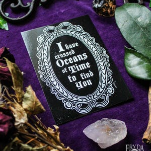 Puede incluir: Una tarjeta negra con un marco blanco adornado y el texto "He cruzado oc&eacute;anos de tiempo para encontrarte". La tarjeta est&aacute; sobre una superficie de terciopelo morado con una llave, un cristal y flores secas.