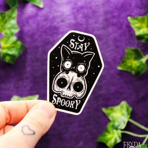 May include: A black and white sticker featuring a cat and skull design with the words "Stay Spooky". The sticker is held by a hand with a heart tattoo. The background is purple with green leaves.