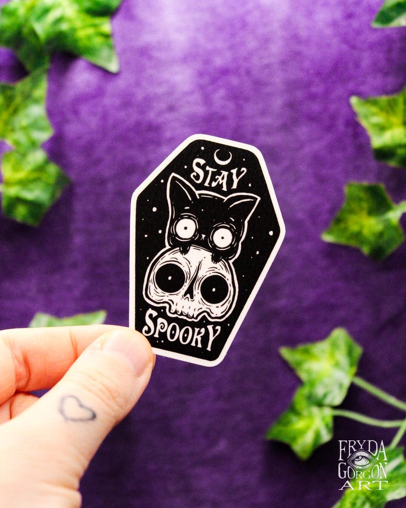 May include: A black and white sticker featuring a cat and skull design with the words "Stay Spooky". The sticker is held by a hand with a heart tattoo. The background is purple with green leaves.