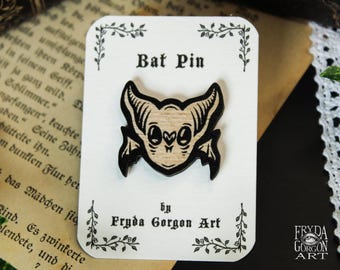 May include: A wooden bat pin featuring a light tan face with black engraved details, large eyes, and pointed ears. The pin is attached to a white rectangular display card with the text Bat Pin and by Fryda Gorgon Art printed in black.