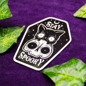 May include: A black and white sticker featuring a cat and skull design with the words "Stay Spooky". The sticker has a white border and is set against a purple background with green leaves.
