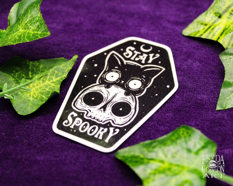 May include: A black and white sticker featuring a cat and skull design with the words "Stay Spooky". The sticker has a white border and is set against a purple background with green leaves.