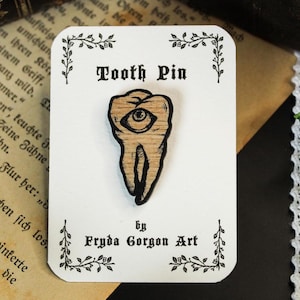 Peut inclure: Épingle en bois en forme de dent avec un œil, présentée sur une carte blanche. L'épingle présente une illustration détaillée d'une dent avec un œil au centre. La carte est ornée d'éléments floraux noirs et du texte "Tooth Pin" et "by Fryda Gorgon Art."