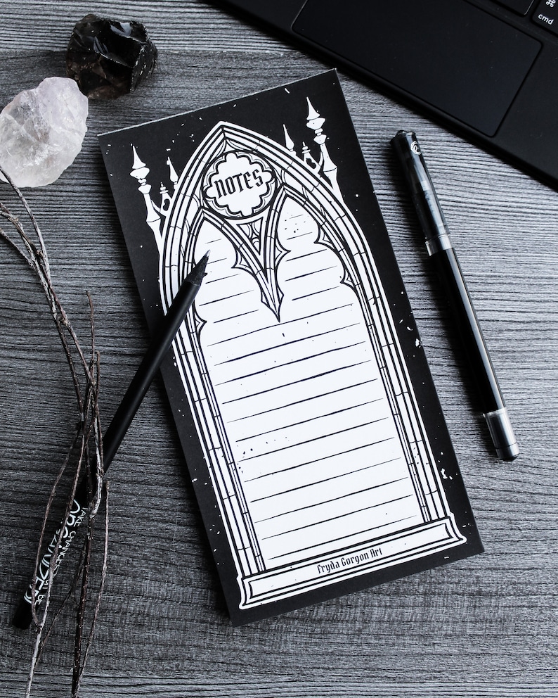 May include: Black and white notepad with a gothic arch design. The notepad has lined pages and the text "notes" in a circle at the top. The bottom of the notepad says "Fryda Gorgon Art".