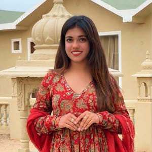 May include: A red floral print dress with long sleeves and a V-neck. The dress features a matching red scarf and is adorned with a floral pattern in shades of green and gold. The woman is wearing gold rings.