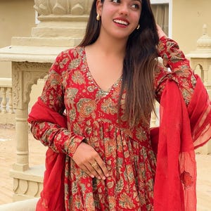 May include: A red floral print dress with long sleeves and a V-neck. The dress has a flowing silhouette and is paired with a matching red scarf. The floral pattern includes shades of green and gold.
