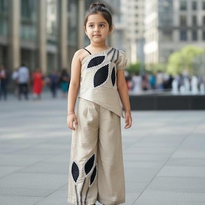 May include: A young girl in a beige two-piece outfit with black leaf-shaped details. The top is asymmetrical with one shoulder covered and a leaf design. The wide-leg trousers have matching leaf details. She wears black sandals.