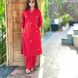 Könnte beinhalten: Ein rotes Tunika- und Hosen-Set. Die Tunika hat einen V-Ausschnitt und weiße Stickereien. Die Person trägt eine Sonnenbrille und gemusterte Ballerinas. Das Outfit ist aus leichtem Stoff gefertigt.