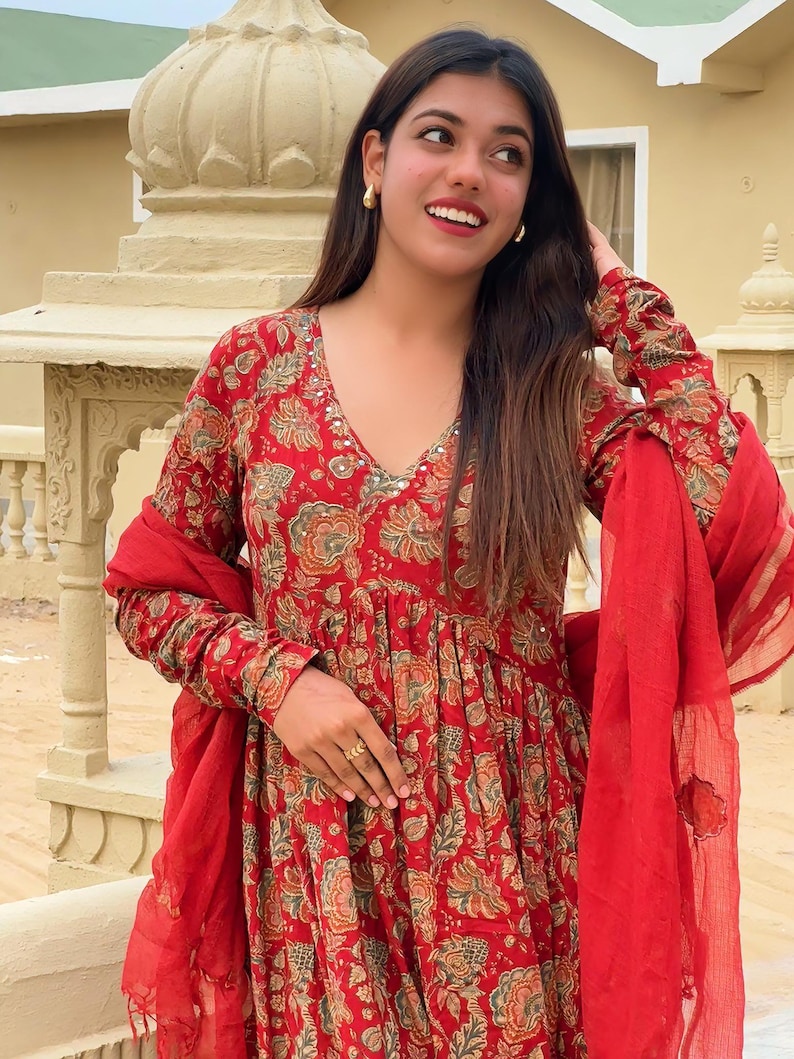 May include: A red floral print dress with long sleeves and a V-neck. The dress has a flowing silhouette and is paired with a matching red scarf. The floral pattern includes shades of green and gold.