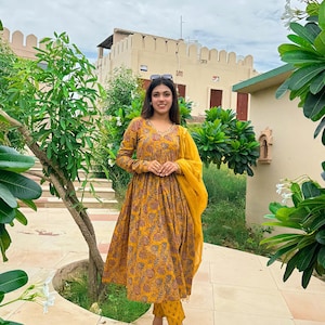May include: A mustard yellow floral print dress with matching pants and a sheer scarf. The long-sleeved dress has a V-neck and a flared skirt. The outfit is worn with clear sandals.