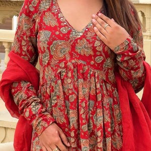 May include: A red floral print dress with long sleeves and a V-neckline. The dress features a gathered waist and is paired with a matching red scarf. The floral pattern includes shades of brown and green.