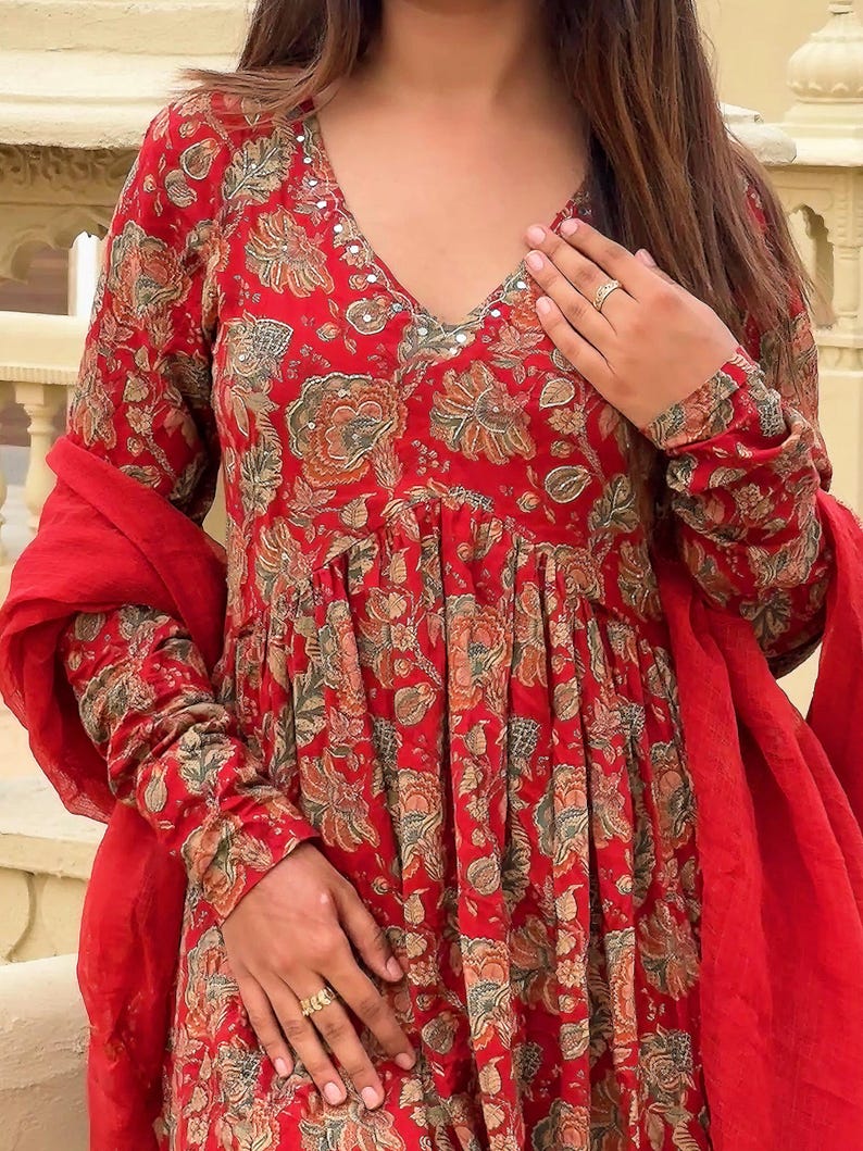 May include: A red floral print dress with long sleeves and a V-neckline. The dress features a gathered waist and is paired with a matching red scarf. The floral pattern includes shades of brown and green.