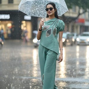 Peut inclure: Une personne porte un haut asymétrique vert clair et un pantalon assorti, tenant un parapluie transparent à motifs floraux blancs. La tenue présente des ornements en forme de feuilles. La personne porte des lunettes de soleil noires et des talons noirs.
