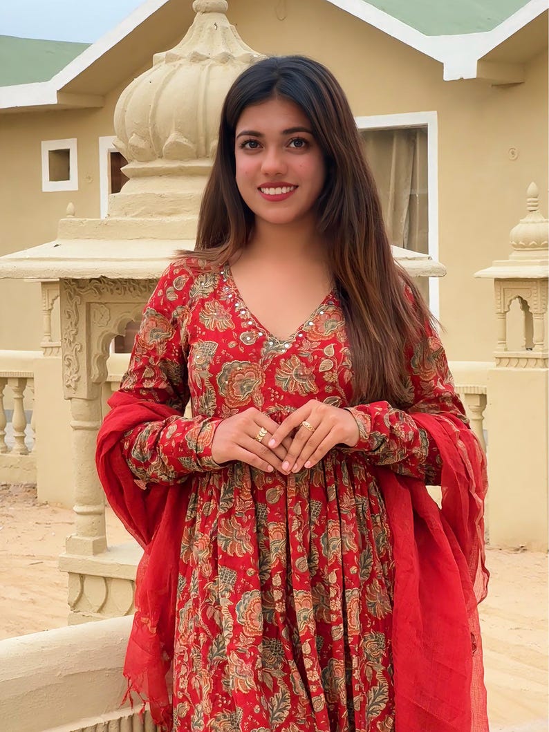 May include: A red floral print dress with long sleeves and a V-neck. The dress features a matching red scarf and is adorned with a floral pattern in shades of green and gold. The woman is wearing gold rings.