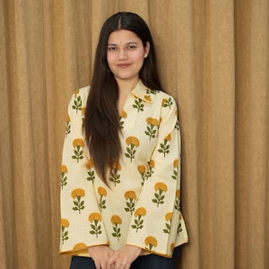 May include: A cream-coloured, long-sleeved top featuring a floral pattern of yellow flowers and green leaves. The top has a collared neckline and is worn with dark blue jeans. The floral print is repeated across the fabric.