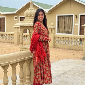 May include: A woman wearing a red and gold floral print dress with matching pants and a red scarf. The dress has long sleeves and a tiered skirt. She is wearing beige flats with gold accents. The background includes a beige building and a stone railing.