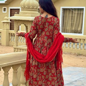 May include: A red floral print salwar kameez set. The long-sleeved tunic and matching pants feature a detailed floral pattern in shades of beige and green. A red scarf is draped over the shoulders. The outfit is a traditional Indian style.