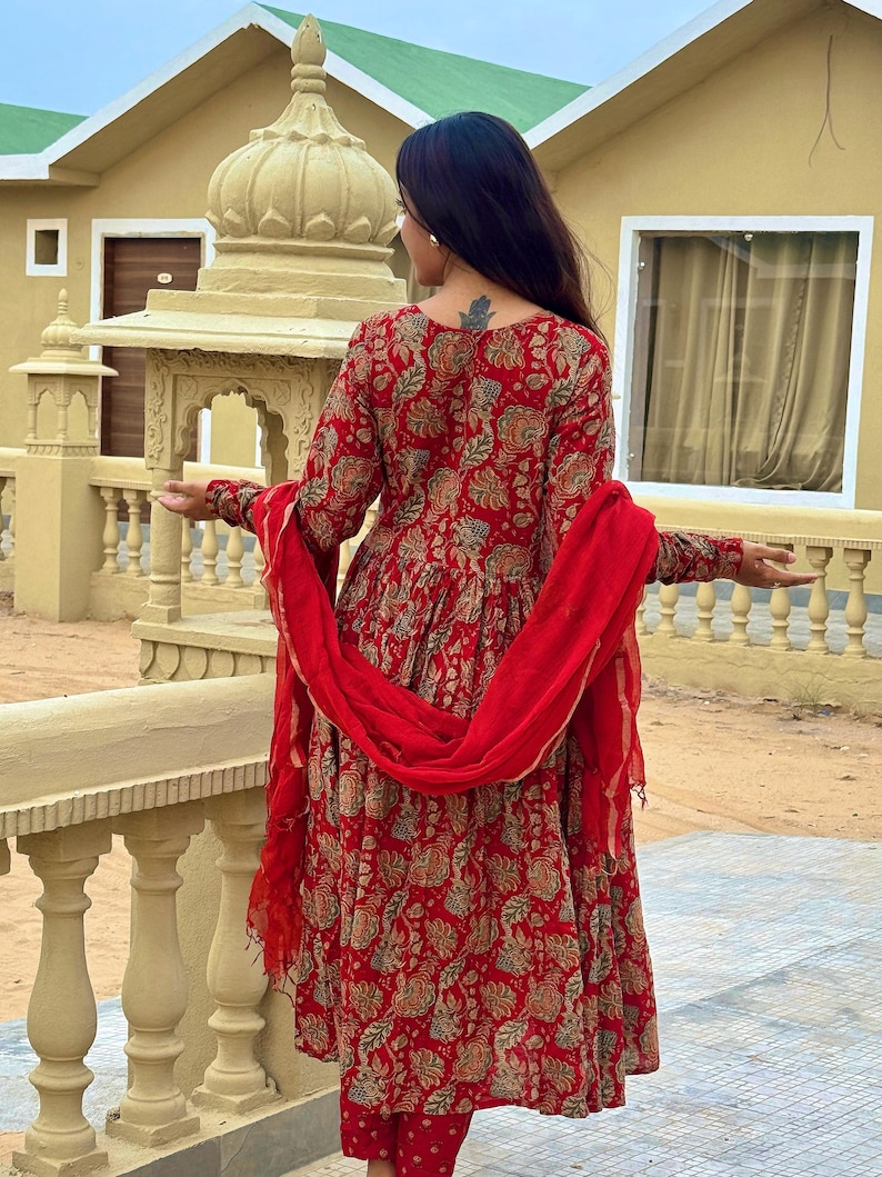 May include: A red floral print salwar kameez set. The long-sleeved tunic and matching pants feature a detailed floral pattern in shades of beige and green. A red scarf is draped over the shoulders. The outfit is a traditional Indian style.
