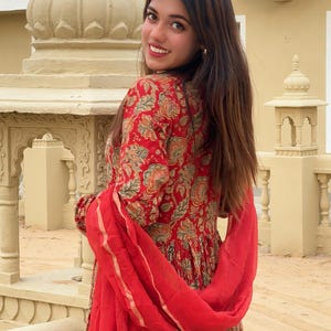 May include: A red floral print dress with a matching red scarf. The dress features a detailed pattern in shades of green, orange, and beige. The scarf is draped over the shoulders, adding a pop of color to the ensemble. The dress has long sleeves.