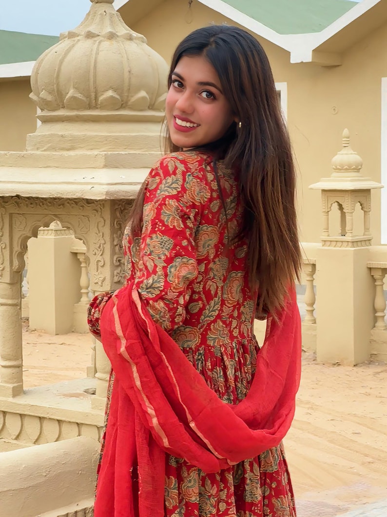 May include: A red floral print dress with a matching red scarf. The dress features a detailed pattern in shades of green, orange, and beige. The scarf is draped over the shoulders, adding a pop of color to the ensemble. The dress has long sleeves.