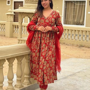 May include: A red floral print Anarkali suit with matching pants and a red dupatta. The long-sleeved dress has a V-neck and is paired with cream-colored shoes. The outfit is suitable for festive occasions.