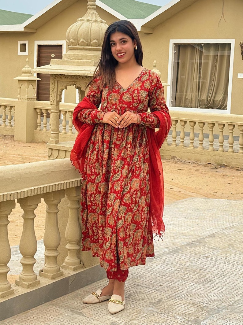 May include: A red floral print Anarkali suit with matching pants and a red dupatta. The long-sleeved dress has a V-neck and is paired with cream-colored shoes. The outfit is suitable for festive occasions.
