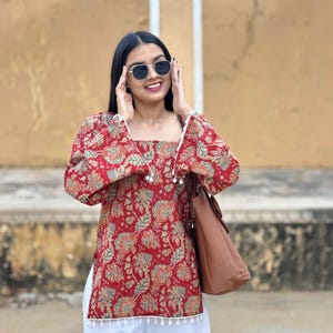 Puede incluir: Una mujer con un top rojo con estampado floral, pantalones blancos y un bolso marrón. El top tiene un escote cuadrado y mangas largas. El estampado floral incluye tonos de rojo, verde y beige. Los pantalones son blancos y de pierna ancha.