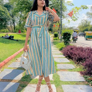 May include: A striped midi dress with green, yellow, and white vertical stripes. The dress has short sleeves, a collared neckline, and a matching belt. The person is wearing white strappy heels and carrying a white handbag.