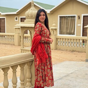 May include: A woman wearing a red and gold floral print dress with matching pants and a red scarf. The dress has long sleeves and a tiered skirt. She is wearing beige flats with gold accents. The background includes a beige building and a stone railing.