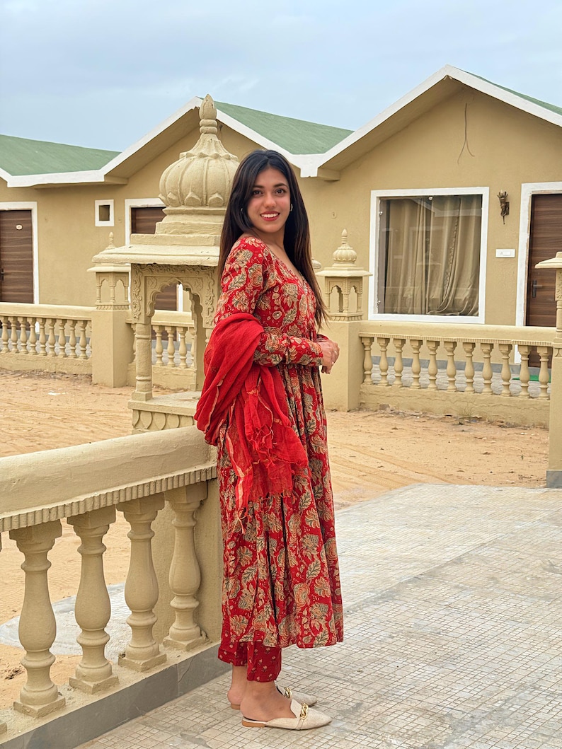 May include: A woman wearing a red and gold floral print dress with matching pants and a red scarf. The dress has long sleeves and a tiered skirt. She is wearing beige flats with gold accents. The background includes a beige building and a stone railing.