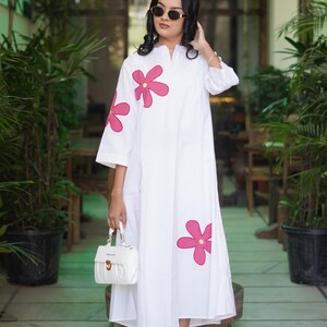 May include: White dress with pink flower accents. The dress has a v-neck and three-quarter sleeves. A white handbag and strappy sandals complete the look. The background features greenery and a cafe entrance.
