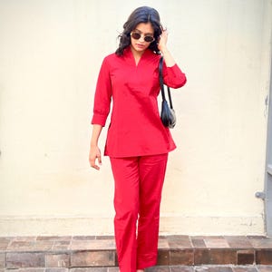 Könnte beinhalten: Ein leuchtend rotes zweiteiliges Outfit, bestehend aus einem Tunika-Oberteil mit Dreiviertelärmeln und einer passenden Hose mit geradem Bein. Das Outfit wird mit gemusterten Ballerinas und einer schwarzen Umhängetasche kombiniert. Die Person trägt eine Sonnenbrille.