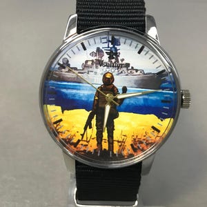 May include: A round wristwatch with a black nylon band. The watch face features a soldier standing on a yellow and blue landscape, with a ship in the background. The watch has silver-colored casing.