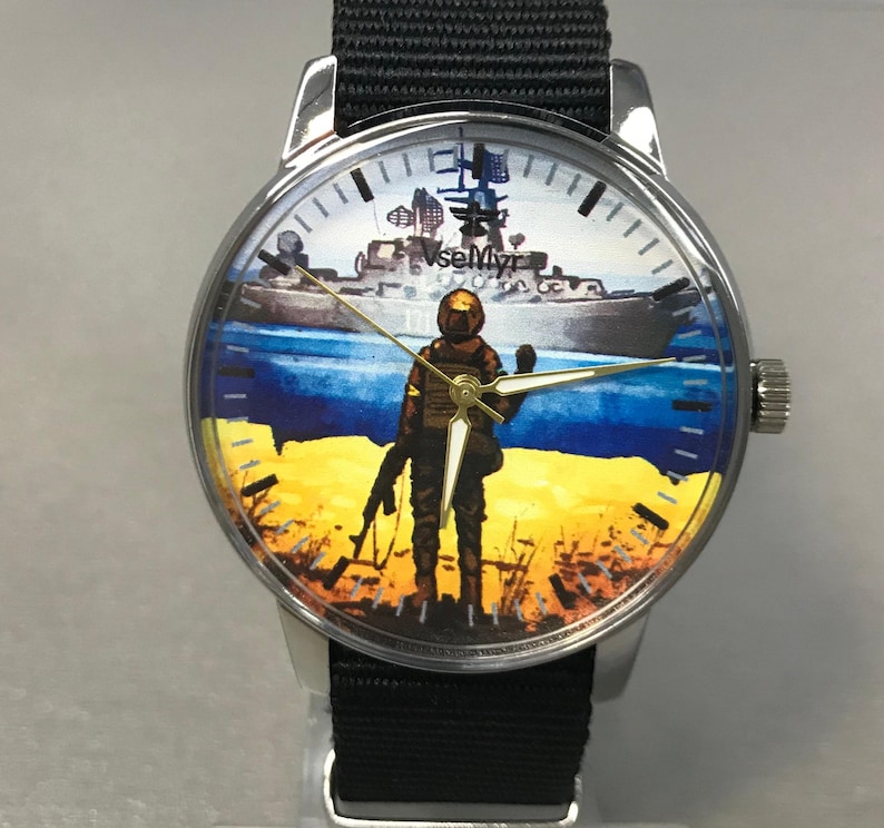 May include: A round wristwatch with a black nylon band. The watch face features a soldier standing on a yellow and blue landscape, with a ship in the background. The watch has silver-colored casing.