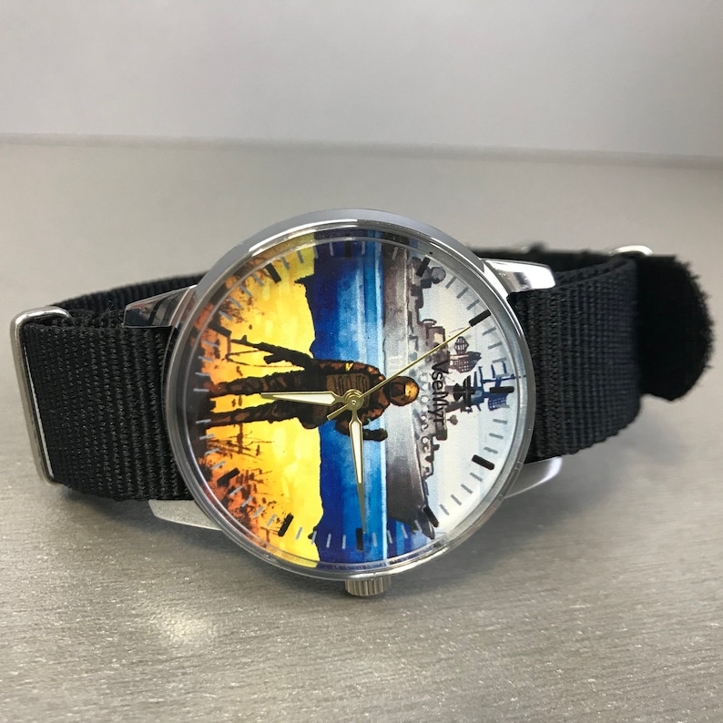 May include: A silver-toned wristwatch with a black nylon strap. The watch face features a split design: one side depicts a figure against a yellow and blue backdrop, the other a cityscape. The watch hands are gold.
